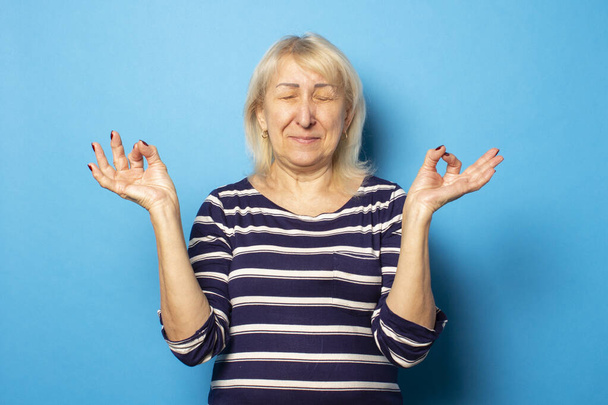 Retrato de una anciana amigable con camiseta casual meditando sobre un fondo azul aislado. Cara emocional. El concepto de relajación, separación
. - Foto, imagen