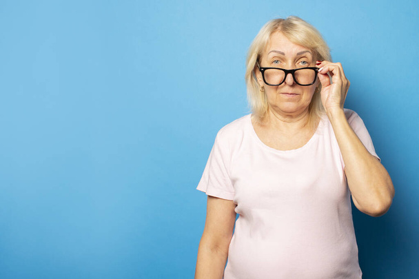 Retrato de uma velha mulher amigável em t-shirt casual e óculos segurando óculos em um fundo azul isolado. Cara emocional. Gesto copos abaixados
. - Foto, Imagem