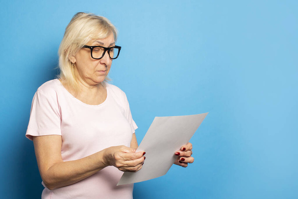 Portrait of an old friendly woman in a casual T-shirt and glasses holding a sheet of paper in her hands and looking at it on an isolated blue background. Emotional face. Concept letter, notice. - Zdjęcie, obraz