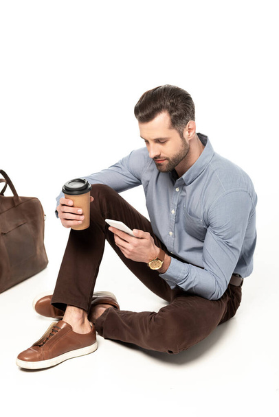 bearded businessman using smartphone while sitting near bag on white  - Foto, afbeelding