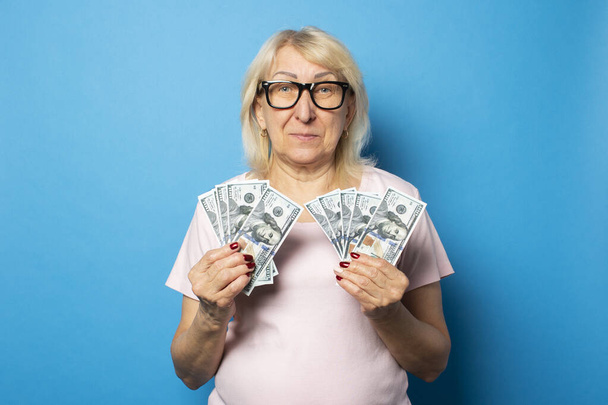 Portrait of an old friendly woman in casual t-shirt and glasses holding money in her hands on an isolated blue background. Emotional face. Concept wealth, win, credit, pension. - Photo, image