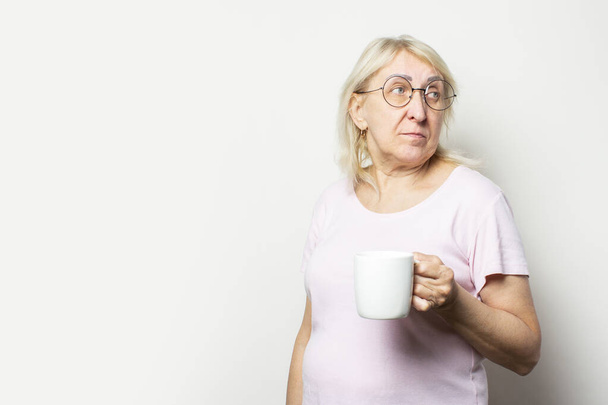 カジュアルなTシャツやメガネを着た笑顔の優しい老女の肖像画は、彼女の手にマグカップを持っており、孤立した光の背景を振り返ります。感情的な顔だ。朝のコーヒーコンセプト、朝食. - 写真・画像