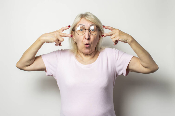 Ritratto di una vecchia donna amichevole con un volto sorpreso in una t-shirt casual e occhiali fa un gesto di benvenuto su uno sfondo leggero isolato
. - Foto, immagini