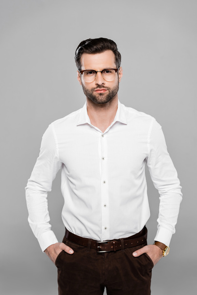 bearded businessman in glasses standing with hands in pockets isolated on grey  - 写真・画像