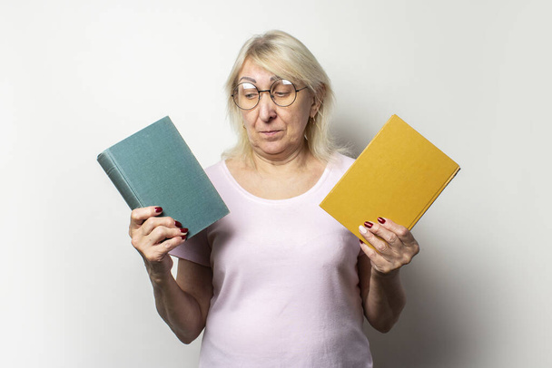 Portrait of an old friendly woman with a smile in a casual t-shirt and glasses holds two books on an isolated light background. Emotional face. Concept book club, leisure, choice of books. - Photo, Image