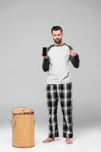 bearded man pointing with finger at smartphone with blank screen near laundry basket on grey  - Фото, зображення
