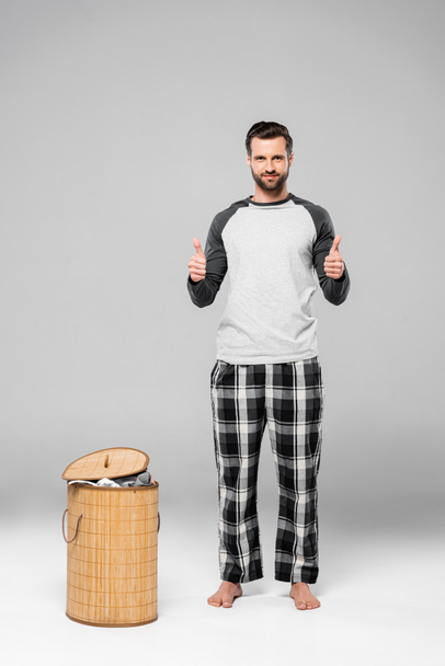 happy man showing thumbs up while standing near laundry basket on grey  - Foto, Imagem