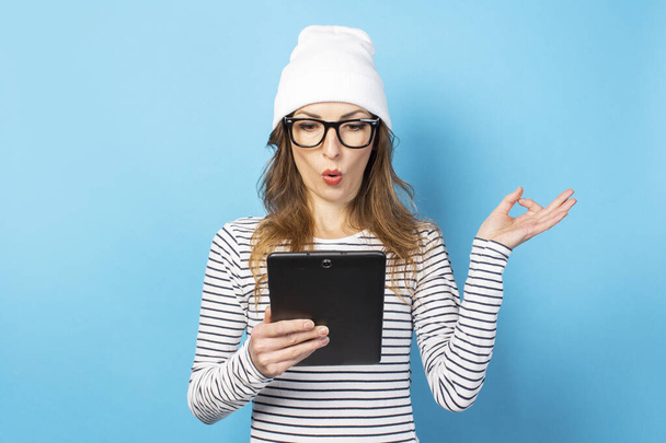 Retrato de una joven amigable con una cara sorprendida en un sombrero, gafas y una camiseta sosteniendo una tableta en sus manos y haciendo un gesto zen sobre un fondo azul aislado. Cara emocional
. - Foto, Imagen