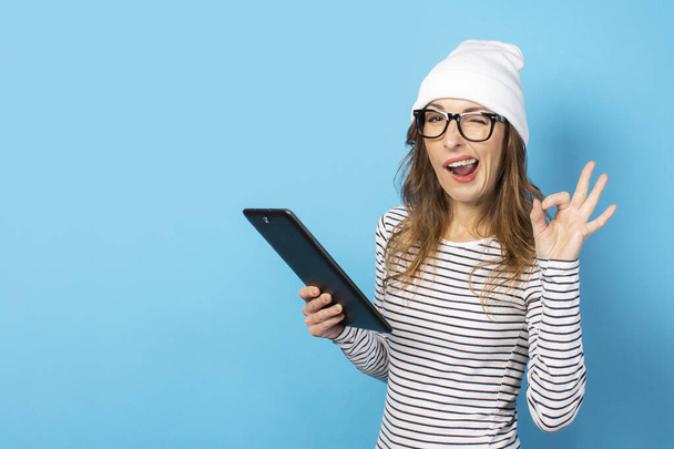 Retrato de una joven amigable con una cara alegre en un sombrero, gafas y una camiseta sostiene una tableta en sus manos y hace el signo OK sobre un fondo azul aislado. Cara emocional
. - Foto, Imagen