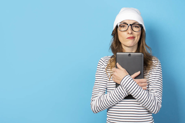 Porträt einer jungen, freundlichen Frau mit verlegenem Gesicht, Hut, Brille und T-Shirt, die auf einem isolierten blauen Hintergrund ein Tablet in der Hand hält. Emotionales Gesicht. - Foto, Bild