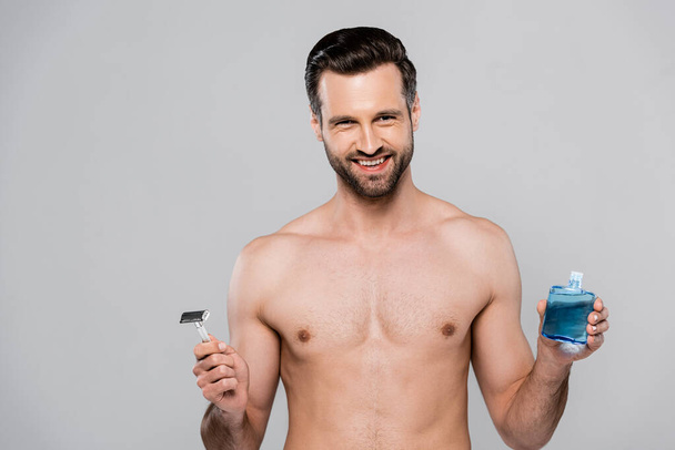 cheerful and muscular man holding after shave lotion and razor isolated on grey  - Fotografie, Obrázek