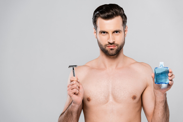 bearded and muscular man holding after shave lotion and razor isolated on grey  - Foto, Imagen