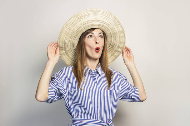 Πορτρέτο μιας νεαρής φιλικής γυναίκας με ένα έκπληκτο πρόσωπο σε ένα φόρεμα και ένα πλατύ καπέλο σε ένα απομονωμένο φόντο φωτός. Συναισθηματικό πρόσωπο. Χειρονομία του αιφνιδιασμού, χαρά. - Φωτογραφία, εικόνα