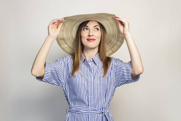 Πορτρέτο μιας νεαρής φιλικής γυναίκας με ένα χαμόγελο σε ένα φόρεμα και ένα πλατύ καπέλο κοιτάζει πάνω σε ένα απομονωμένο φόντο φως. Το καπέλο καλύπτει μέρος του προσώπου. Συναισθηματικό πρόσωπο. Χειρονομία χαράς. - Φωτογραφία, εικόνα