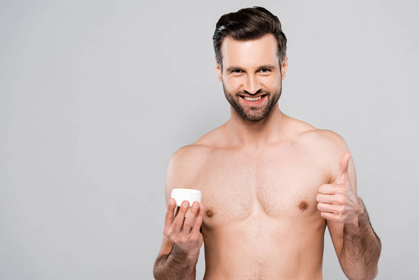 happy bearded man holding container with cosmetic cream and showing thumb up isolated on grey  - Foto, Imagem