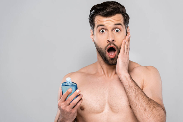 šokovaný muž drží láhev s po holení krém a dotýká tvář izolované na šedé  - Fotografie, Obrázek