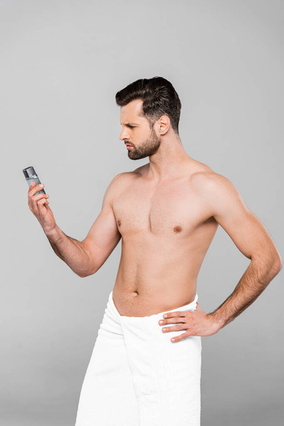 bearded and muscular man in towel looking at deodorant while standing with hand on hip isolated on grey  - Foto, Bild
