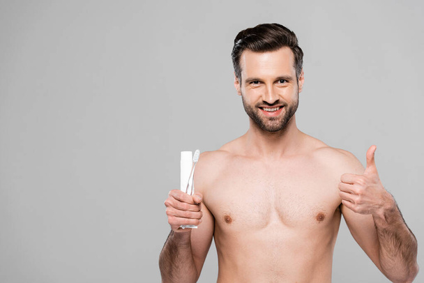 happy and muscular man holding toothpaste and toothbrush while showing thumb up isolated on grey  - Φωτογραφία, εικόνα