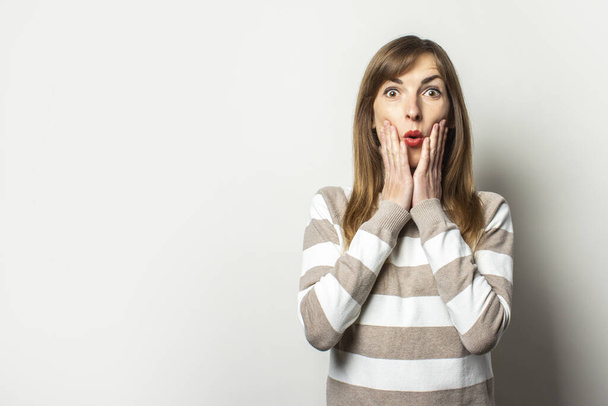 Portrait of a young friendly woman in a striped sweater and a surprised face on an isolated light background. Emotional face. Gesture surprise shock. - Photo, Image
