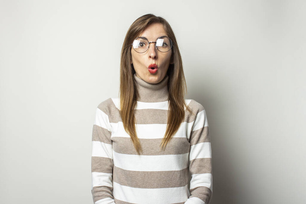 Retrato de uma jovem mulher amigável em uma camisola listrada e óculos com um rosto surpreso em um fundo leve isolado. Cara emocional. Choque surpresa de gestos
. - Foto, Imagem