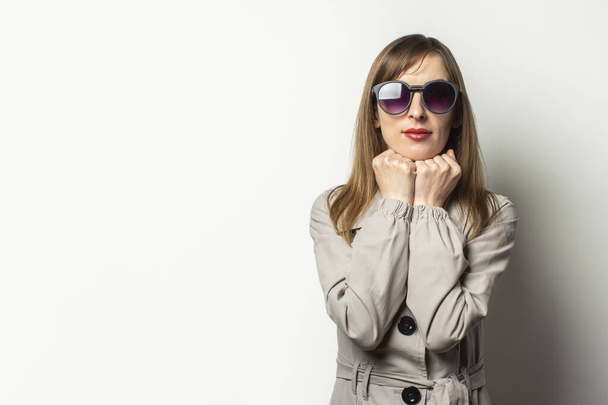 Portrait of a young friendly woman in a classic jacket and sunglasses holding her hands under her chin and smiling on an isolated light background. Emotional face. Classic style concept, french woman. - Photo, Image