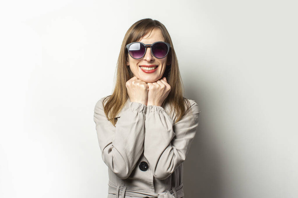 Portrait of a young friendly woman in a classic jacket and sunglasses holding her hands under her chin and smiling on an isolated light background. Emotional face. Classic style concept, french woman. - Fotoğraf, Görsel