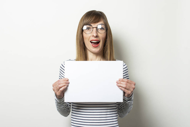 Portrait of a young friendly woman in a long-sleeve sweater and glasses with a surprised face holds a blank sheet of paper on light background. Emotional face. Gesture shock, surprise. Copy space. - Foto, imagen