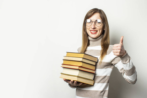 Young friendly woman in sweater and glasses holding stack of books in her hands and makes gesture Everything is fine, thumb up on light background. Emotional face. Education concept, exam preparation. - Photo, Image