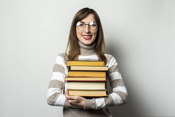 Πορτρέτο μιας νεαρής φιλικής γυναίκας με πουλόβερ και γυαλιά που κρατά μια στοίβα βιβλία στα χέρια της σε ένα απομονωμένο φόντο φωτός. Συναισθηματικό πρόσωπο. Εκπαίδευση έννοια, προετοιμασία εξετάσεις. - Φωτογραφία, εικόνα