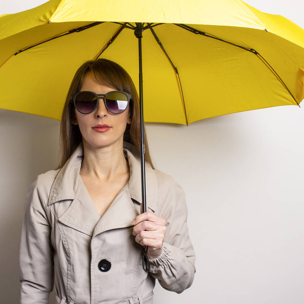Χαρούμενη χαρούμενη νεαρή γυναίκα με μαύρα γυαλιά ηλίου που γελάει κρατά μια κίτρινη ομπρέλα σε γκρι φόντο. Έννοια της βροχής, κακός καιρός. - Φωτογραφία, εικόνα