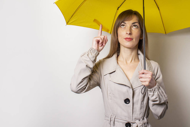 Χαρούμενη χαρούμενη νεαρή γυναίκα κρατά μια κίτρινη ομπρέλα και δείχνει ένα δάχτυλο προς τα πάνω σε ένα γκρι φόντο. Έννοιες χειρονομίες, βροχή, κακός καιρός. - Φωτογραφία, εικόνα