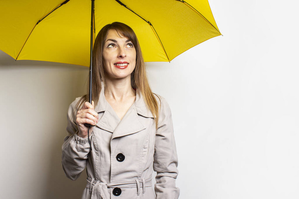 Portré Fiatal barátságos nő klasszikus kabátban, sárga esernyő alatt mosollyal felnéz egy elszigetelt fényháttérre. Érzelmes arc. Rossz idő, eső, időjárás előrejelzés. - Fotó, kép