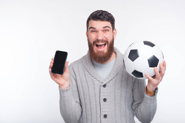 young amazed man holding smartphone and soccer ball over white background - Photo, image