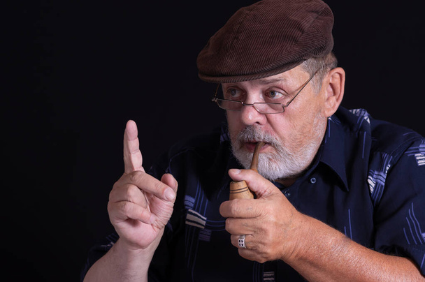 Καλλιτεχνικό πορτραίτο γενειοφόρου ηλικιωμένου άνδρα που κρατά πίπα καπνού και δείχνει δείκτη σε μαύρο φόντο - Φωτογραφία, εικόνα