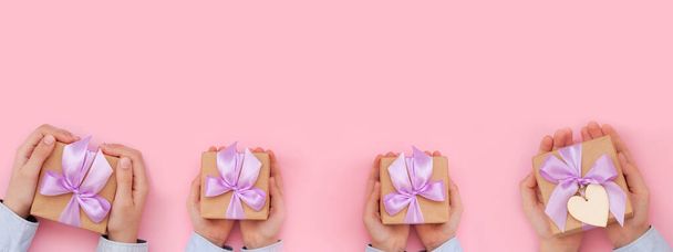 Kid's hands holding gift box wrapped in craft paper and tied with bow on pink background. Concept Mother's Day or Birthday greeting card. Banner - Photo, Image