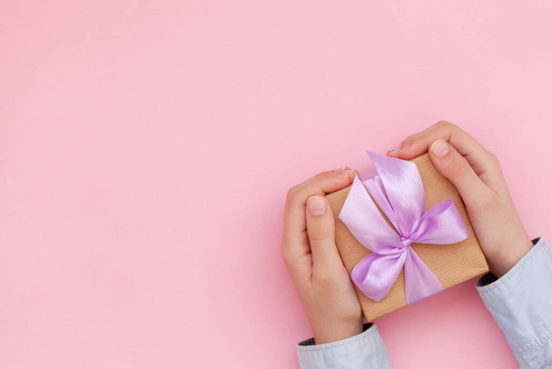 Kid's hands holding gift box wrapped in craft paper and tied with bow on pink background. Concept Mother's Day or Birthday greeting card. - Fotoğraf, Görsel