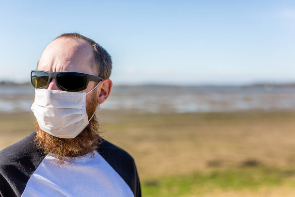 Ένας γενειοφόρος άνδρας με προστατευτική μάσκα προσώπου και γυαλιά ηλίου κατά τη διάρκεια της παγκόσμιας πανδημίας της κορώνας. - Φωτογραφία, εικόνα