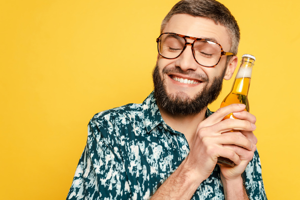smiling bearded guy in glasses with closed eyes and bottle of beer isolated on yellow - Foto, Imagem