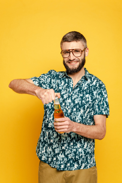 smiling bearded guy in glasses opening bottle of beer isolated on yellow - Foto, afbeelding