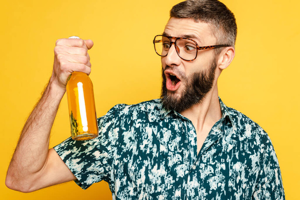 wstrząśnięty brodaty facet w okularach patrząc na butelkę piwa izolowanego na żółto - Zdjęcie, obraz