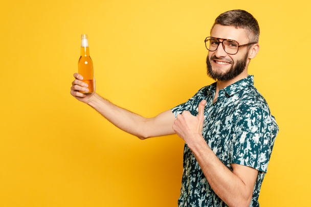 side view of happy bearded guy in glasses with refreshing bottle of beer showing thumb up on yellow - Photo, Image