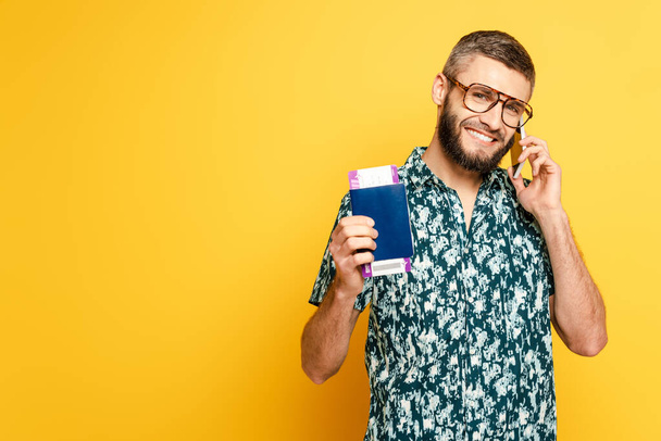 smiling bearded guy in glasses with passport and ticket talking on smartphone on yellow - Foto, Bild