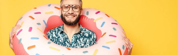 smiling bearded guy in glasses with donut swim ring isolated on yellow,  panoramic orientation - Fotó, kép