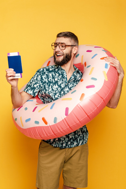 smiling bearded guy in glasses with donut swim ring and passport on yellow - Fotoğraf, Görsel