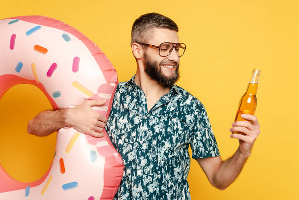 smiling bearded guy in glasses with donut swim ring and beer on yellow - Foto, afbeelding