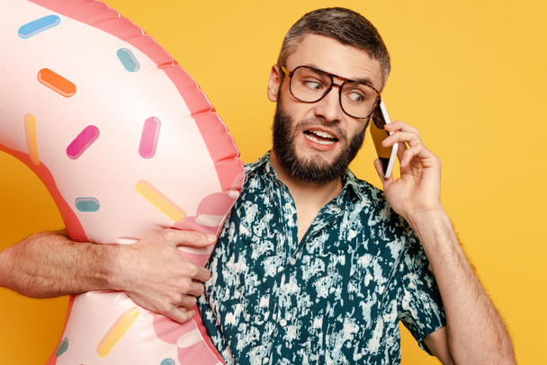 confused bearded guy in glasses with donut swim ring talking on smartphone on yellow - Фото, зображення