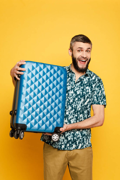 χαρούμενος γενειοφόρος τύπος με μπλε βαλίτσα σε κίτρινο - Φωτογραφία, εικόνα