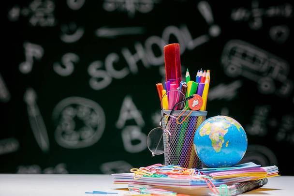School background with stationery accessories. Books, globe, pencils and various office supplies lying on the desk on a green blackboard background. - Photo, Image