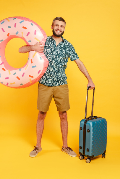 full length view of happy bearded guy with travel bag and swim ring on yellow - Foto, imagen
