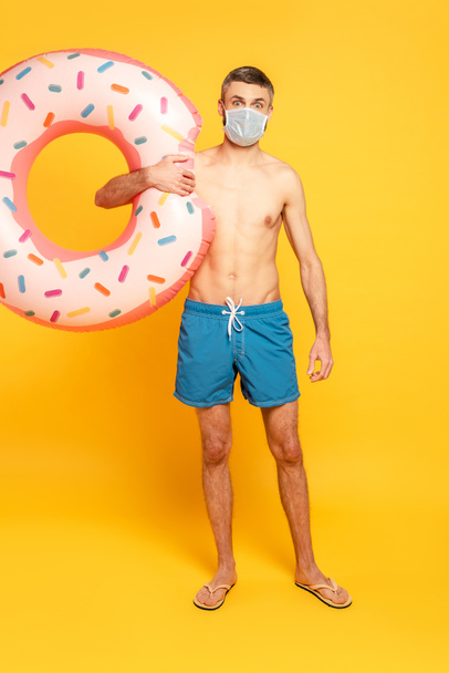 full length view of guy in swim trunks and medical mask with swim ring on yellow - Zdjęcie, obraz